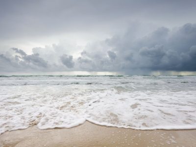 Stormy sea at a sandy beach with foamy waves approaching shore under dark clouds in the sky and a moody horizon.