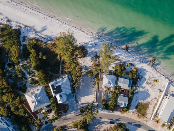 Aerial view of a beachfront neighborhood: white sandy beach, turquoise water, and palm trees surrounding modern houses along a curved street.