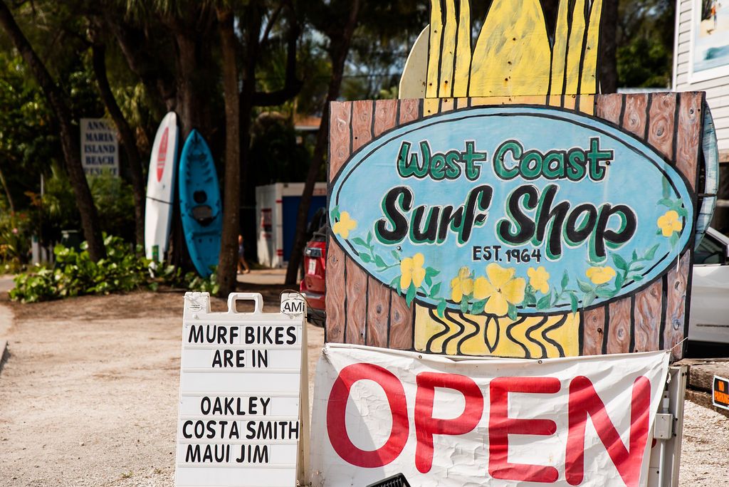 Colorful surf shop sign reading 'West Coast Surf Shop' with an 'OPEN' banner in front at a beachside storefront.