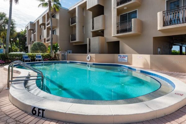 Oval-shaped outdoor pool with turquoise water, beige deck, and palm trees beside a multi-story apartment building.