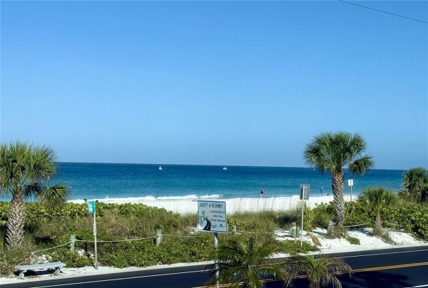 Sunny beach scene with white sand, turquoise ocean, and palm trees; a roadside sign reads 'Adopt-A-Highway'.