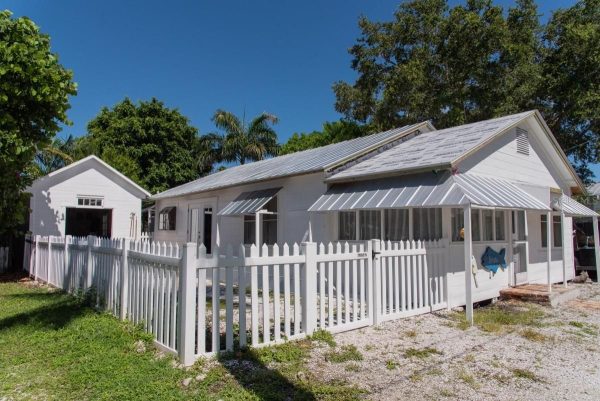 White, single-story coastal cottage with a metal roof, enclosed by a white picket fence and a small covered porch under a bright blue sky.