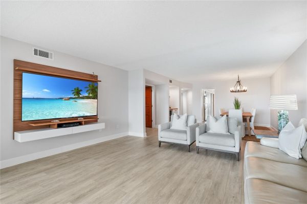 Open-concept living room with a wall-mounted flat-screen TV on a wooden panel, white shelf, and light gray seating area.