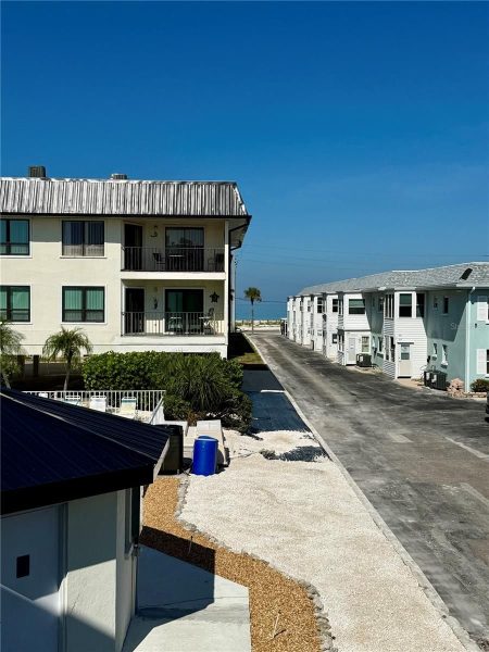 Coastal street with a two-story beige building on the left and a row of white beach houses on the right, leading to the ocean under a clear blue sky.