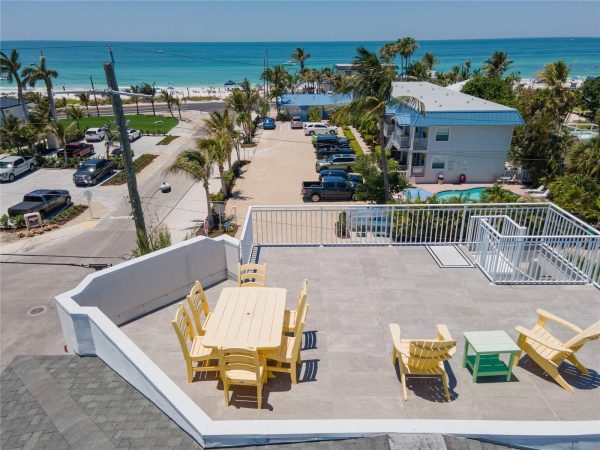 Aerial view of a seaside town with a rooftop patio and yellow outdoor tables and chairs overlooking palm trees, street, and turquoise ocean.