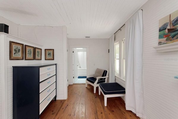 Bright white-paneled hallway with hardwood floor, a navy dresser, four botanical prints, and a seating nook with a chair and ottoman near a window.