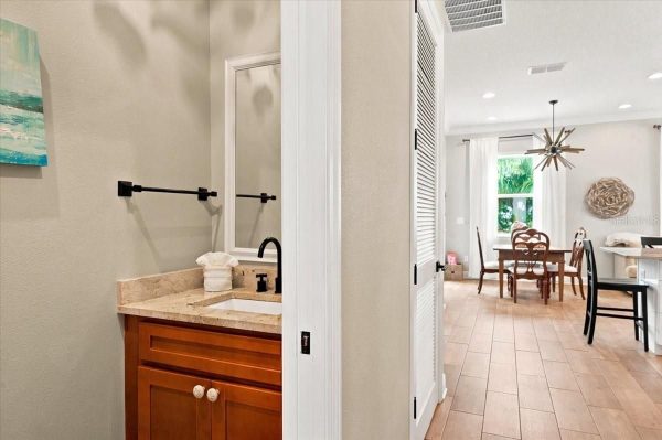 Open-concept hallway: bathroom vanity on the left with beige countertop and black faucet, leading to a dining area with a wooden table and starburst chandelier.