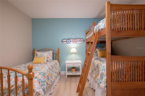 Bedroom with a blue accent wall, two single beds and a wooden bunk bed, white nightstand, lamp, and a surf’s up sign above the table.