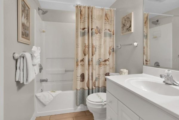 Bright bathroom with tub and seashell-pattern shower curtain, white vanity, toilet, and towels on a wall rack.
