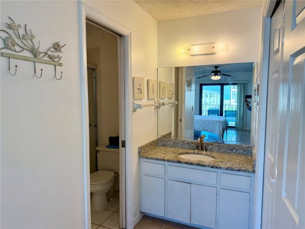 White bathroom vanity with granite countertop and single sink, large mirror above, and door to a bedroom with balcony view reflected in the mirror