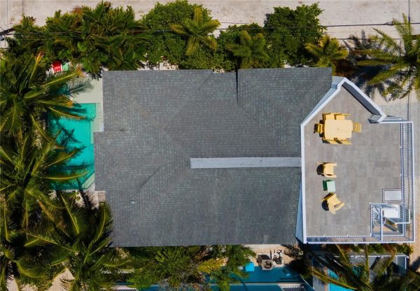 Aerial view of a gray-shingled house roof, turquoise pool at left, and a rooftop terrace with yellow chairs on the right.
