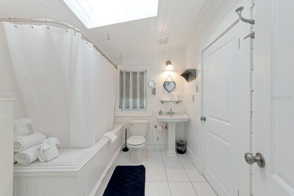 Small white bathroom with a bathtub, shower curtain, toilet, and pedestal sink under a skylight, bright and airy.