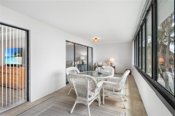Enclosed sunroom with a round glass dining table and white wicker chairs, bright and airy with palm trees outside the large windows.