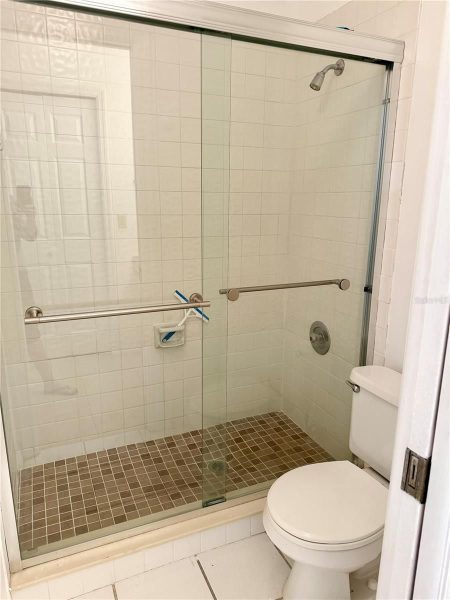 Glass sliding-shower enclosure in a white-tiled bathroom with a brown tile floor and a nearby toilet; two grab bars on the walls and a toothbrush holder visible in a niche.