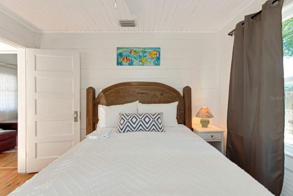 White-paneled bedroom with a wooden bed, white quilt, and a navy geometric throw pillow, topped by a seaside painting above the headboard.