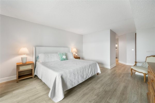 Bright, modern hotel-style bedroom with a large white bed, two wooden nightstands with lamps, and light wood flooring.