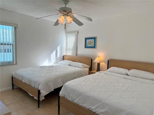 A hotel-style bedroom with two twin beds, white quilts, and woven headboards against white walls, sunlit from a window on the left.