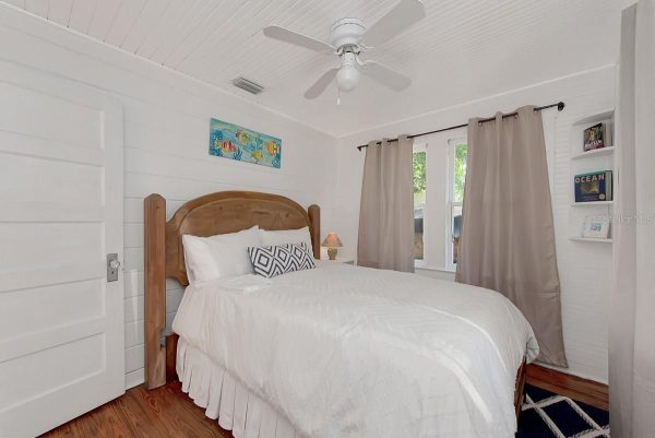 Bedroom with white shiplap walls, wooden bed frame, white bedding, and beige curtains by a window.