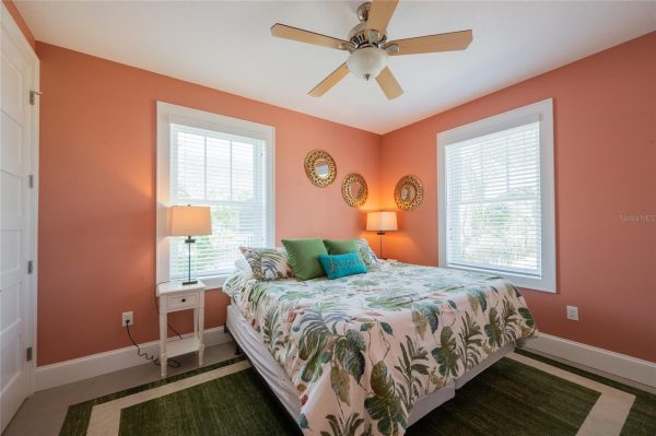 Bedroom with coral walls, queen bed in tropical leaf bedding, two windows, ceiling fan, and mirrored wall decor