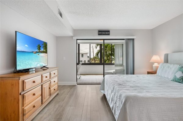 Bedroom with a large bed, nightstand lamp, and wooden dresser with a TV, facing a sliding glass door to a balcony with a tropical view.
