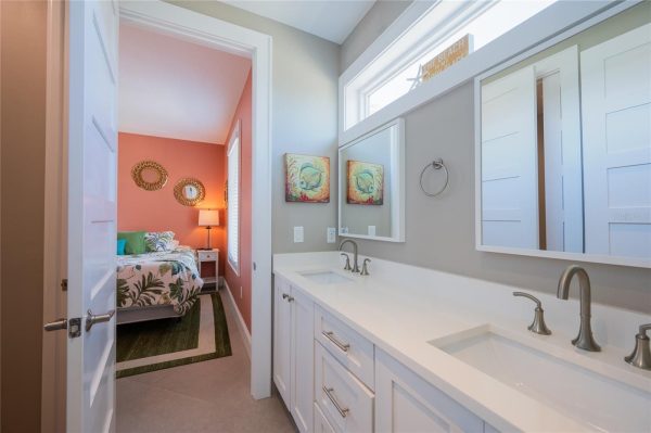 Bathroom with a white double-sink vanity and two mirrors opens to a coral bedroom with tropical bedding and round wicker wall mirrors above a nightstand lamp.