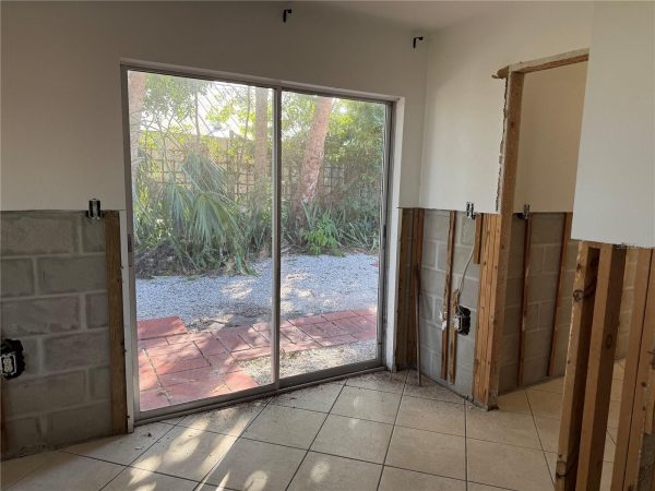 Room under renovation showing exposed studs, a partially removed wall, and a sliding glass door to a garden with palm plants outside.