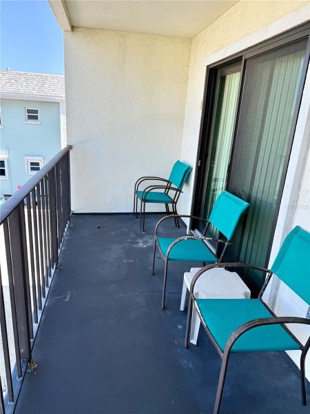 Empty balcony with four turquoise chairs along a sliding glass door and a black railing, neighboring building visible in the distance.