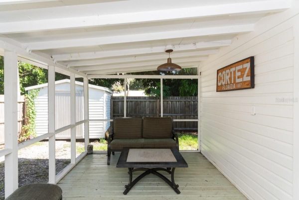 Covered patio with white beams, brown cushioned sofa, and a glass-top coffee table; a Cortez Florida 1887 sign on the wall.