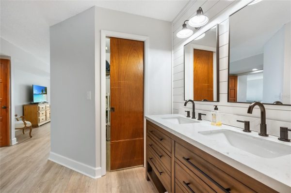 Double-sink bathroom vanity with a white countertop, dark wood drawers, and large mirrors above; doorway to a bedroom visible nearby.