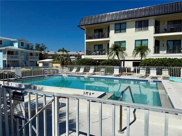 Outdoor pool area at a sunny resort, with white railings, lounge chairs, and multi-story beige buildings in the background under a clear blue sky.