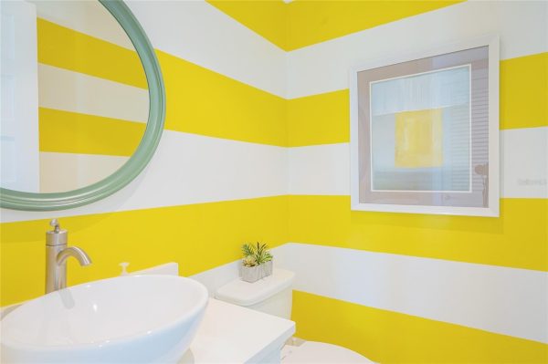 Bright bathroom with wide yellow and white horizontal stripes, round mint-green framed mirror, vessel sink, and a small plant on the toilet tank.