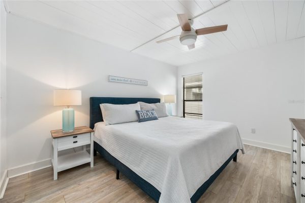 Bright coastal bedroom with a blue-upholstered bed, white linens, two nightstands, and a ceiling fan.