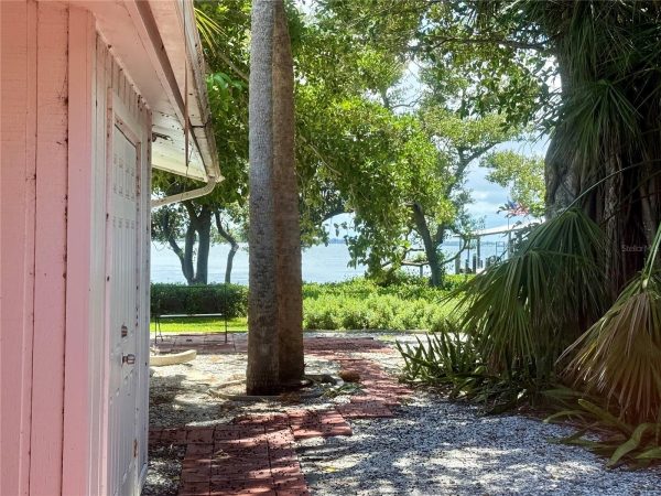 Pink wooden wall and white door on the left, brick pathway, and tropical trees with a water view in the distance.
