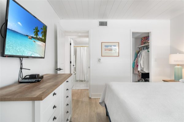 Bright bedroom with a wall-mounted flat-screen TV above a wooden dresser, open closet with clothes, and a neatly made bed to the right.",