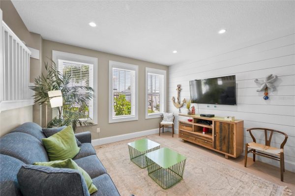 Cozy living room with a blue sofa, green pillows, three tall windows, and a wood TV console beneath a wall-mounted flat-screen TV on a white panel wall.