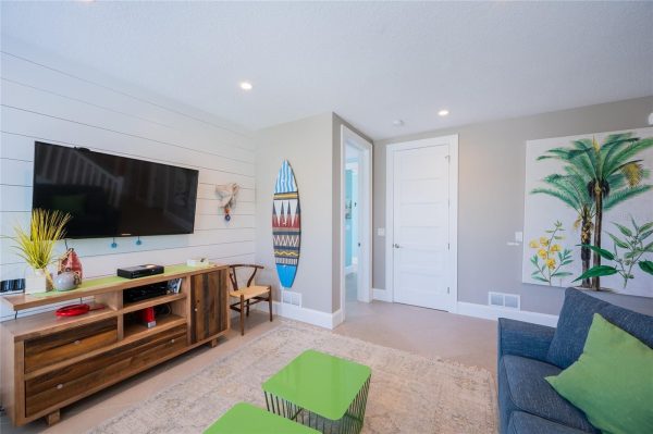 Living room with a wall-mounted TV above a wooden console, decorative objects, and a light gray shiplap wall on the left side; a surfboard wall decor leans against a partition nearby.