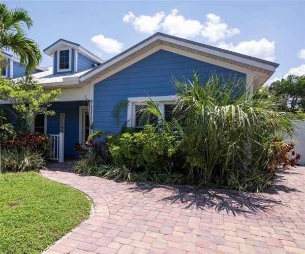 Front view of a blue house with white trim, tropical plants in the yard, and a brick-paver walkway leading to the entrance, under a sunny sky.