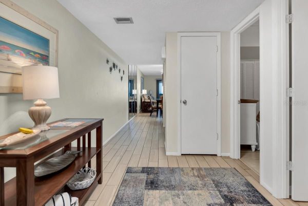 Hallway with a wooden console table, lamp, and beach artwork on the left; doors and a rug extend toward a living area in the distance.