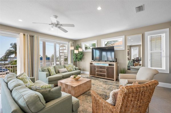 Bright coastal living room with pastel green sofas, a wicker chair, and a TV on a wooden console facing large windows and sliding glass doors to an outdoor area.