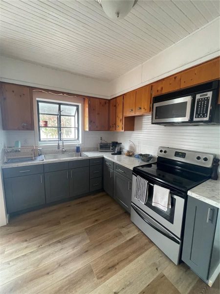 L-shaped kitchen with gray lower cabinets, wooden upper cabinets, stainless stove and microwave, and a window above the sink.