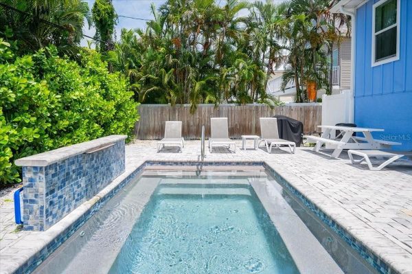 Backyard rectangular pool with blue tiled edge, four lounge chairs, and a wooden privacy fence with tropical plants behind it.