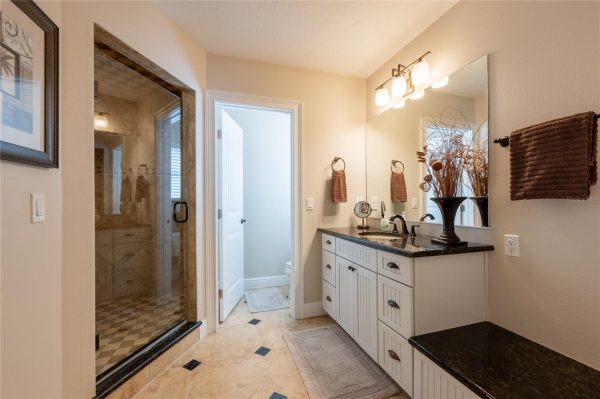 Bathroom with a glass-enclosed shower on the left, a white vanity with a dark countertop, and a large mirror reflecting warm light. A brown towel hangs on the wall and a decorative vase with dried branches sits on the counter. Beige tile flooring with darker inlays.
