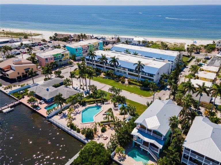 Aerial view of a beachfront town with pastel-colored houses, palm trees, a large white building, and a canal with boat docks beside a pool area by the shore.