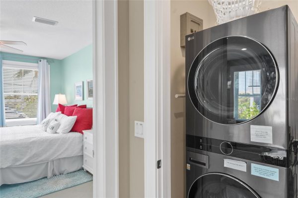 Doorway to a bright bedroom with a white bed and red pillows, seen from a laundry area with a stacked washer and dryer to the right