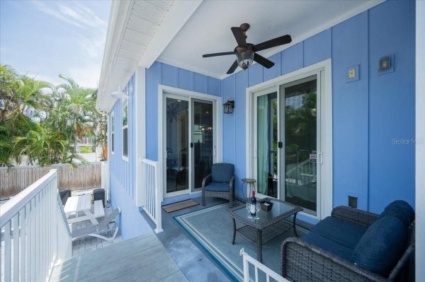 Blue-painted balcony with sliding glass doors, wicker seating, and a glass coffee table on a light rug under a ceiling fan keep this sunny outdoor lounge ready for relaxing.