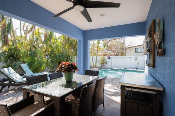 Covered patio with a glass-top dining table and wicker chairs, blue walls, and a ceiling fan overlooking a pool area.