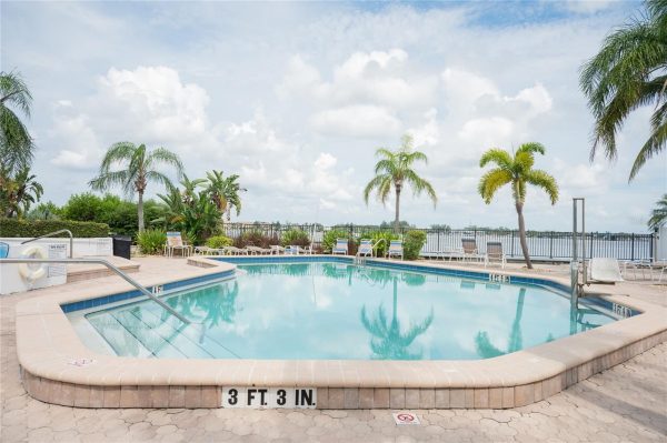 Resort pool surrounded by palm trees and lounge chairs under a blue, partly cloudy sky; railing overlooks water in the distance.