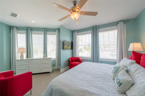 Bright teal bedroom with white furniture, red chairs, and a ceiling fan; large windows with white blinds and light curtains behind a white bedspread.