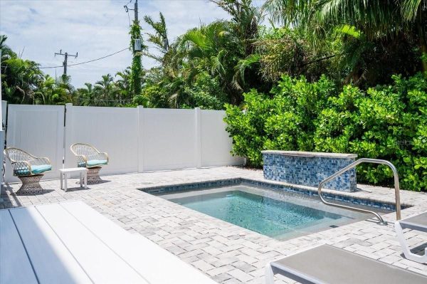Backyard pool area with light-gray pavers, a white privacy fence, two wicker chairs, and tropical greenery behind.