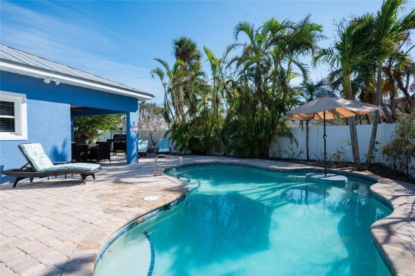 Poolside backyard with a curved turquoise pool, blue house, and tropical palm trees; lounge chair and seating under a beige umbrella on a brick patio.